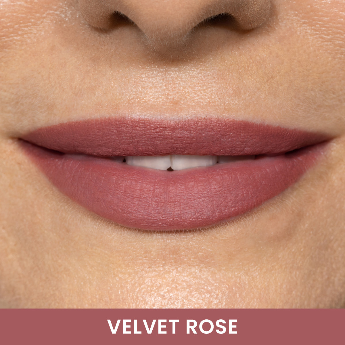 Close-up of a persons lips wearing matte mauve-pink lipstick, labeled Velvet Rose at the bottom. The lips are slightly parted, showing white teeth.