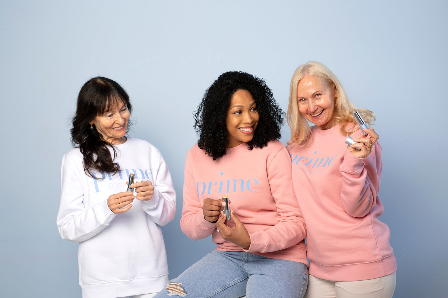 Three women wearing prime sweatshirts smile and hold skincare or beauty products against a light blue background. They appear happy and engaged, showcasing the items to each other.
