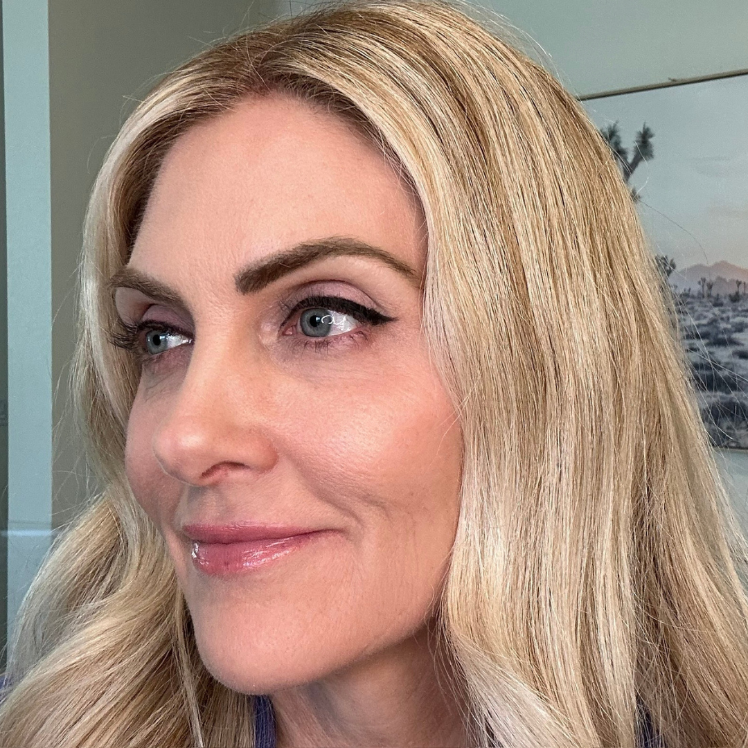 A woman with long, wavy blonde hair and subtle makeup smiles softly while looking to the side; a desert landscape picture hangs on the wall behind her.