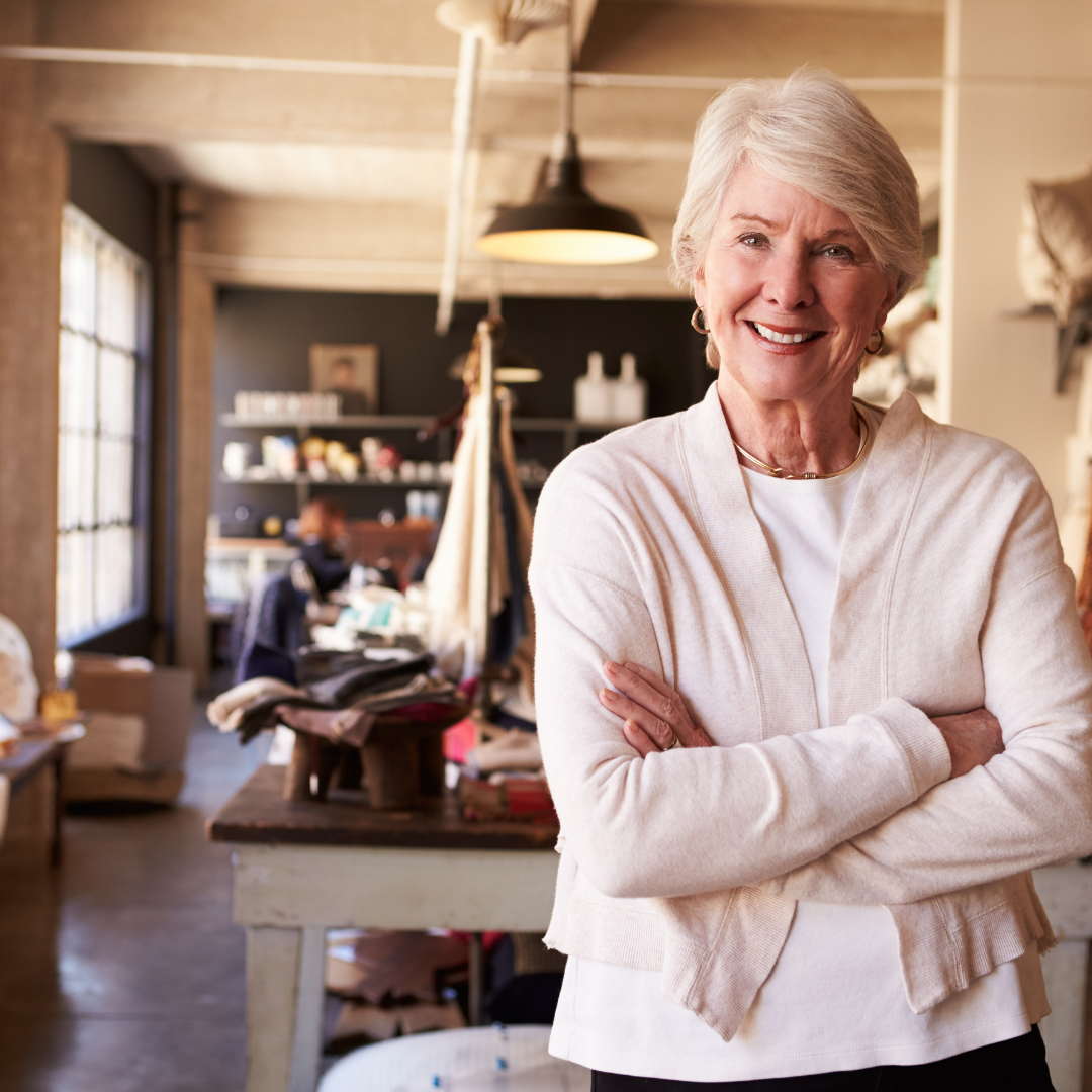 Older woman with short gray hair stands smiling with arms crossed in a bright, modern studio or workshop. The background shows shelves, tables, and people working, suggesting a creative or collaborative environment.
