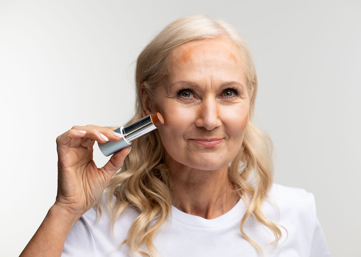 Smiling older woman with long blonde hair applies makeup foundation stick to her forehead and cheeks, wearing a white shirt against a plain light background.