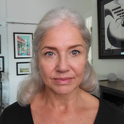A woman with long, gray hair and blue eyes looks directly at the camera, wearing a black top. She is indoors, with framed art and a shelf visible in the background.