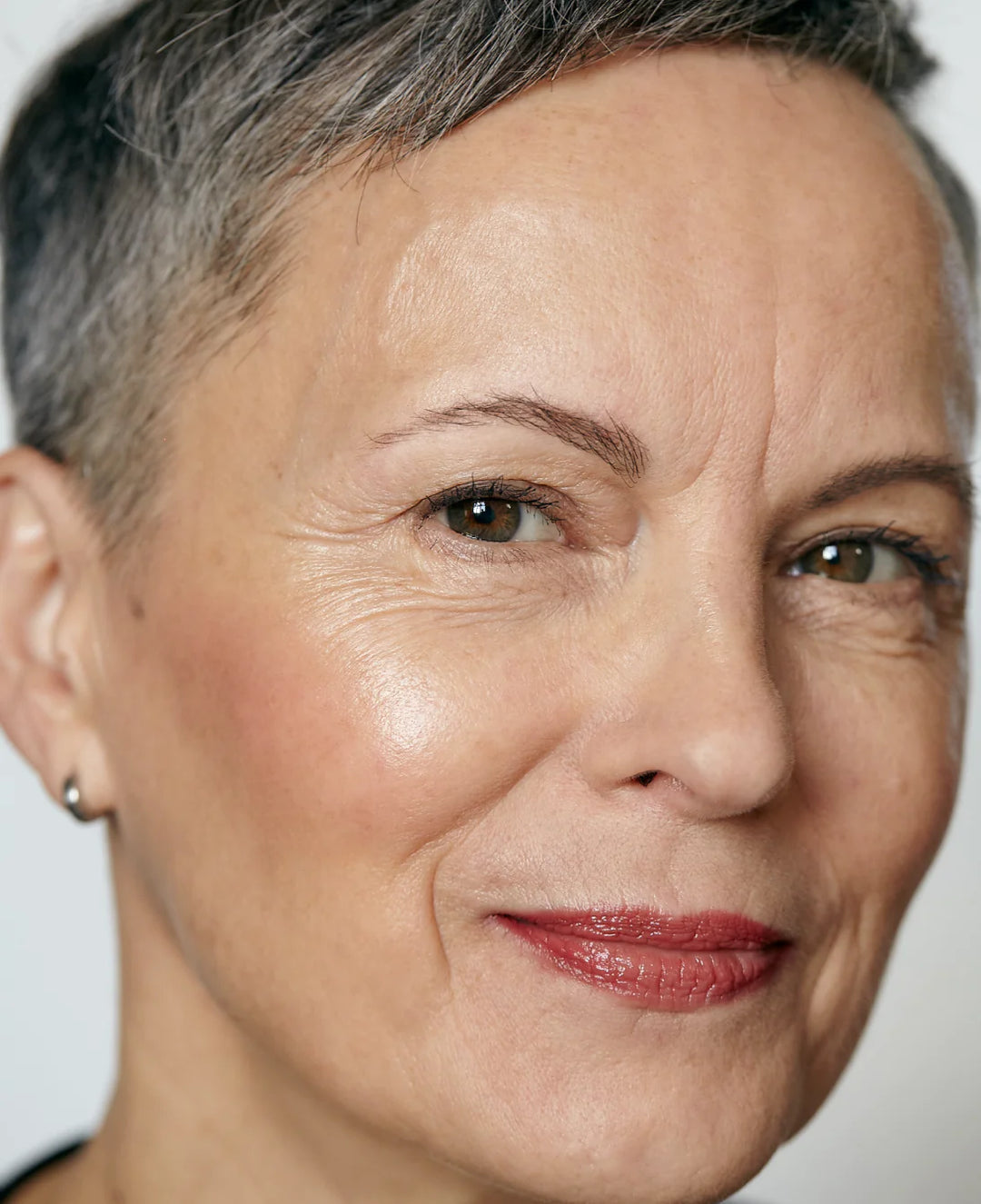 Close-up of an older woman with short gray hair, light skin, and green eyes, smiling softly. She is wearing subtle makeup with red lipstick and a small hoop earring. The background is plain and light-colored.