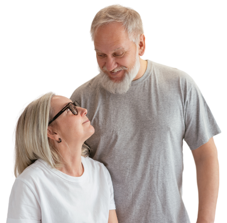 An older man and woman with gray hair smile warmly at each other. The man has a beard and wears a gray t-shirt; the woman wears glasses and a white shirt. Both appear happy and relaxed, standing close together.