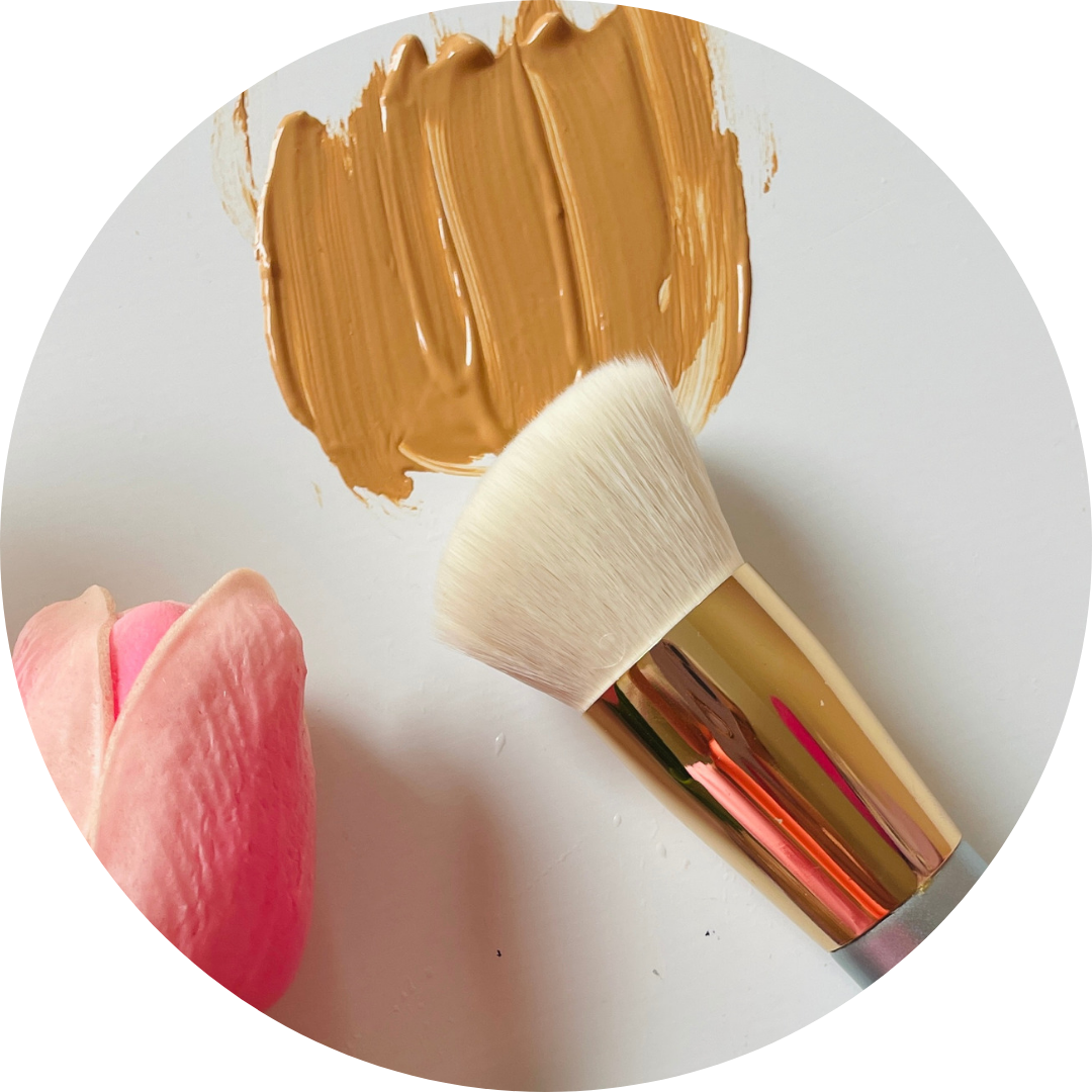A makeup brush with a gold handle lies next to a pink rose petal, with tan foundation smeared on a white surface in the background.