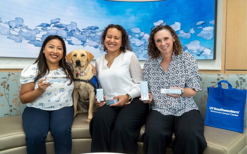 Three women sit on a couch smiling at the camera, holding small boxes. A yellow Labrador in a blue vest sits beside them. A blue UT Southwestern Simmons Cancer Center bag is on the couch to the right.