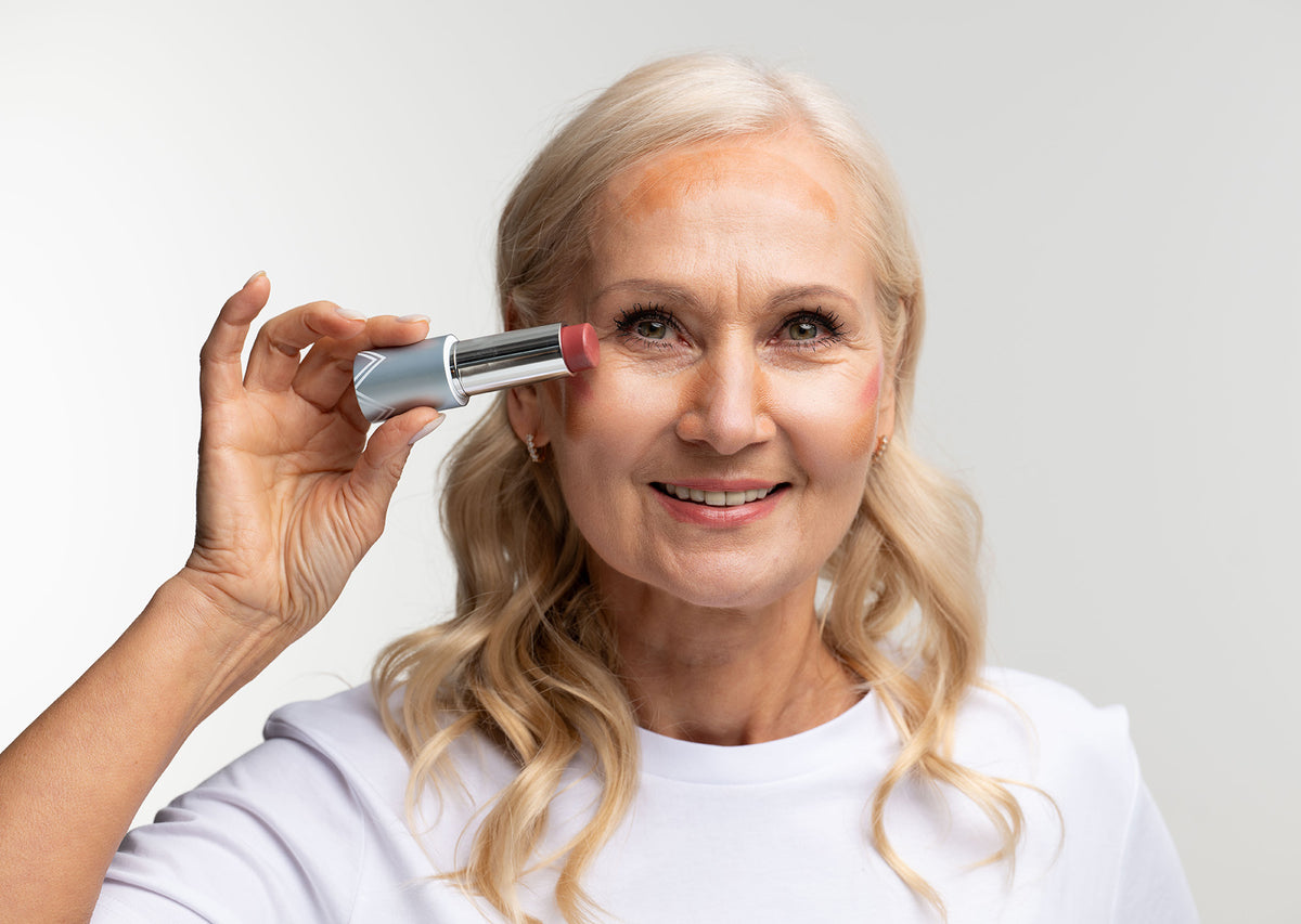 Smiling older woman with long blonde hair holds a tube of lipstick near her face, wearing a white shirt, against a plain light background.