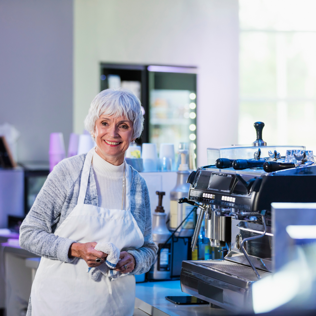 An older woman with short gray hair, wearing a white apron and sweater, smiles while standing beside a commercial coffee machine in a brightly lit café.