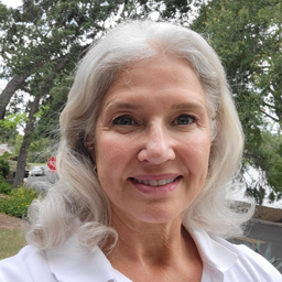 A smiling older woman with wavy gray hair stands outdoors, wearing a white shirt. Trees and greenery are visible in the background.