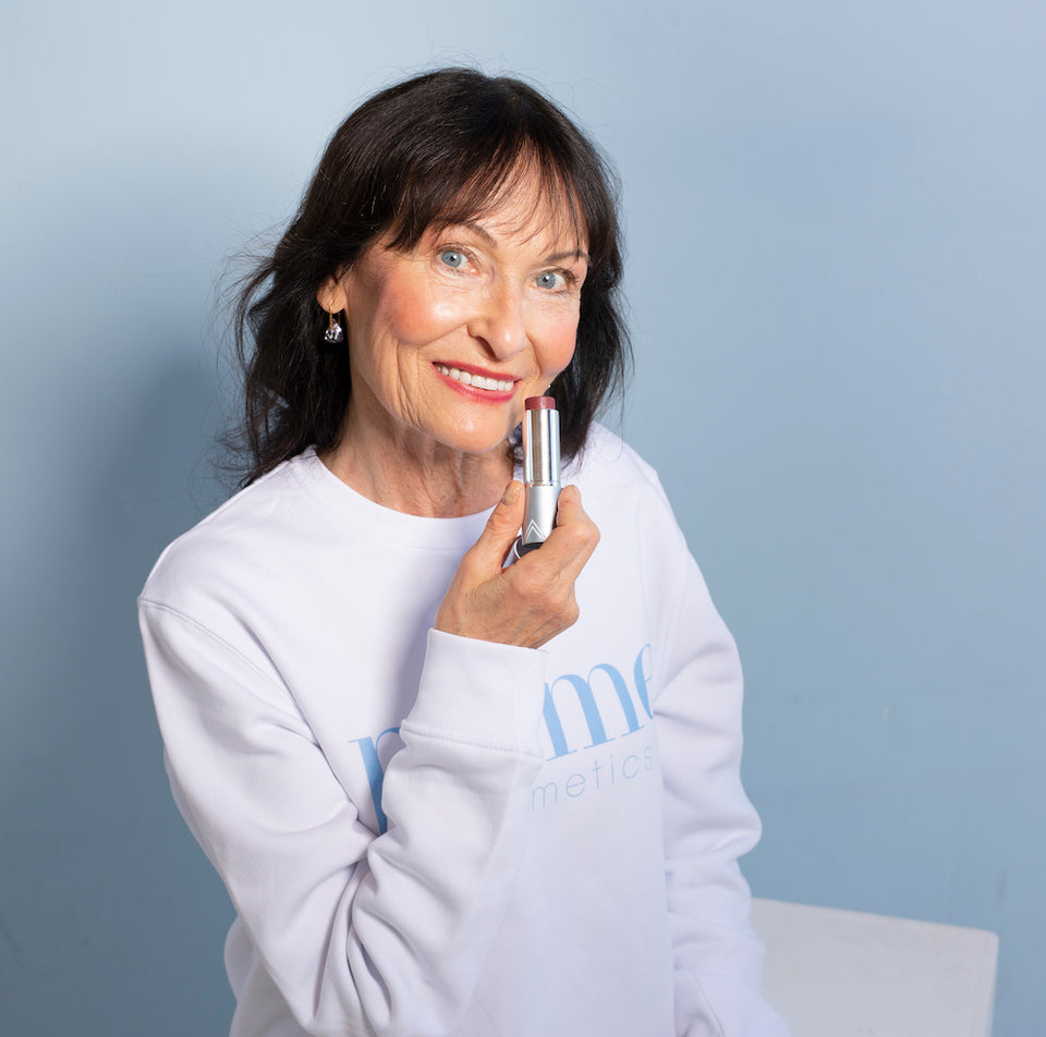 An older woman with long dark hair and blue eyes smiles while holding a lipstick tube to her chin. She is wearing a white long-sleeve shirt and standing against a light blue background.