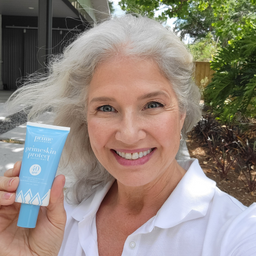 Smiling older woman with long gray hair holding a tube of Prime Primeskin Protect SPF 40 sunscreen outdoors, wearing a white polo shirt, with trees and plants in the background.