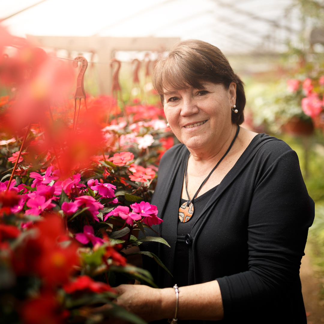 A woman with short brown hair smiles while standing among vibrant pink and red flowers in a greenhouse, wearing a black top and necklace. Sunlight filters softly through the glass roof above.