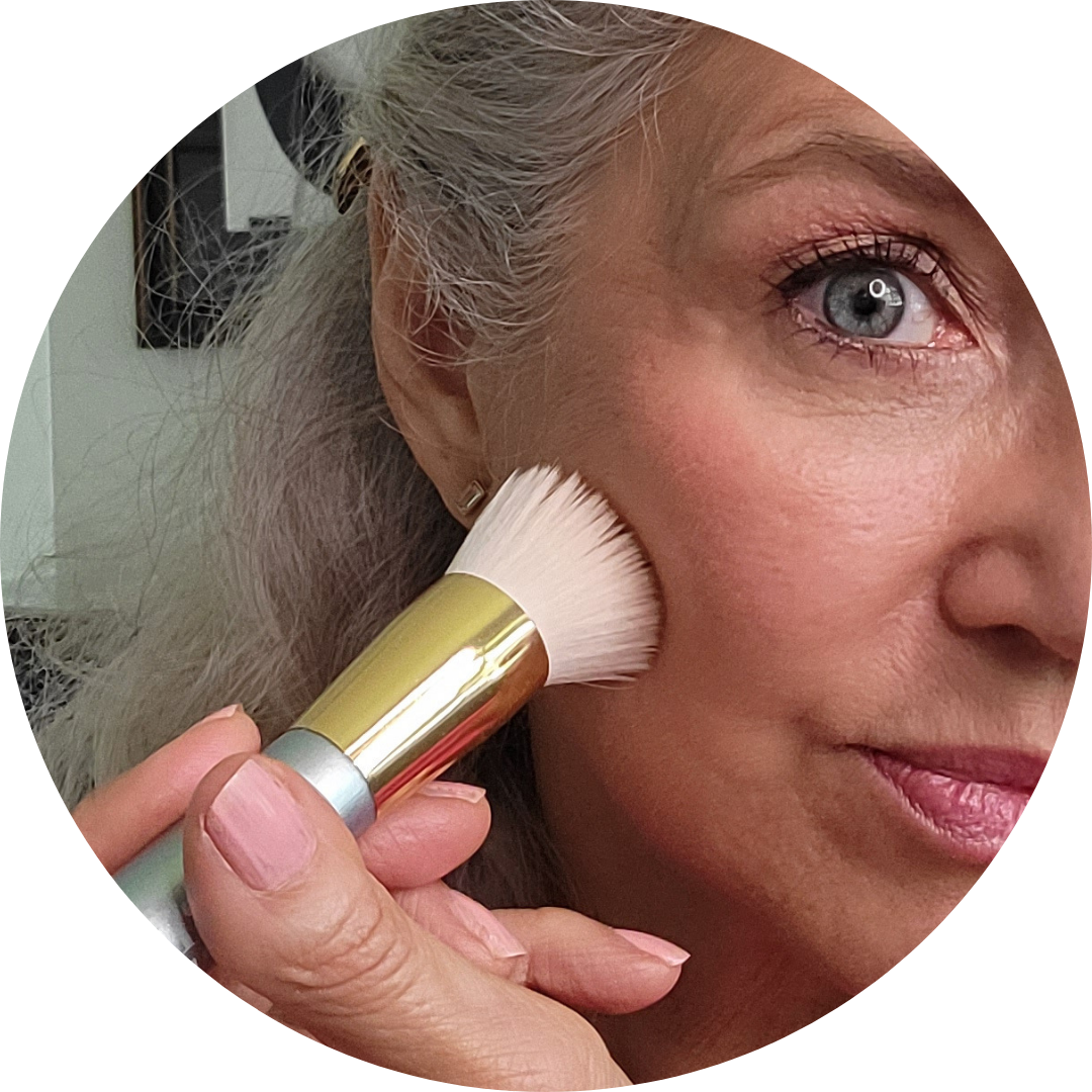 Close-up of a woman with gray hair applying makeup to her cheek with a brush. Her eye and part of her face are visible, showing smooth skin and subtle makeup.