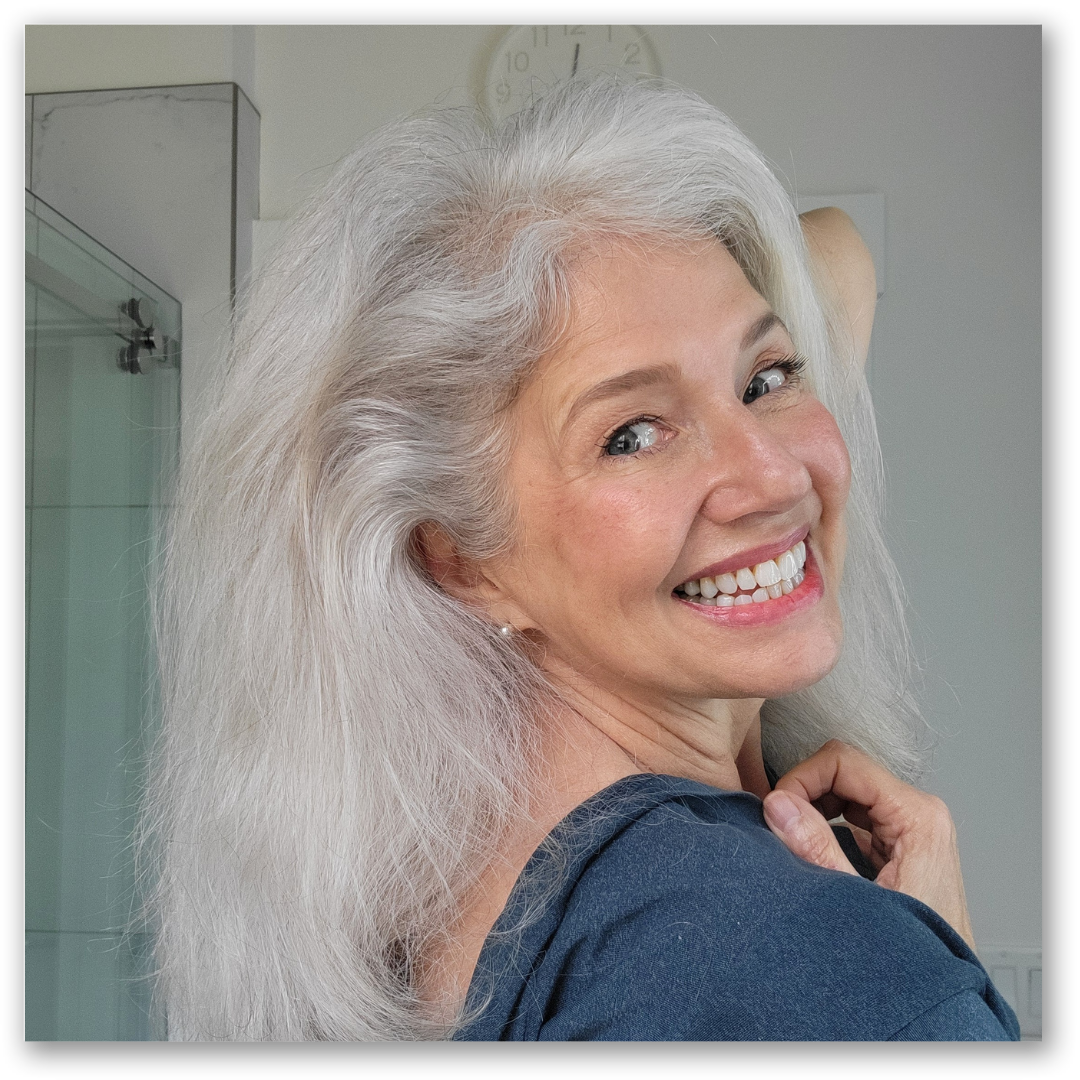 Smiling woman with long, wavy gray hair and blue eyes looking over her shoulder, wearing a dark top, standing in a bright room with a shower and a clock visible in the background.