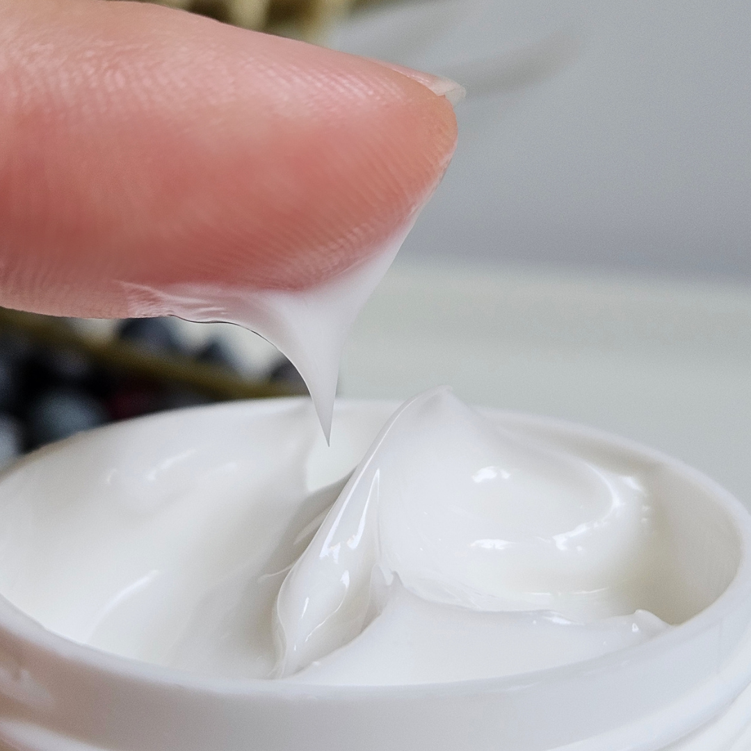A close-up of a finger scooping white cream from an open jar, with the creamy texture stretching from the jar to the fingertip. The background is softly blurred.