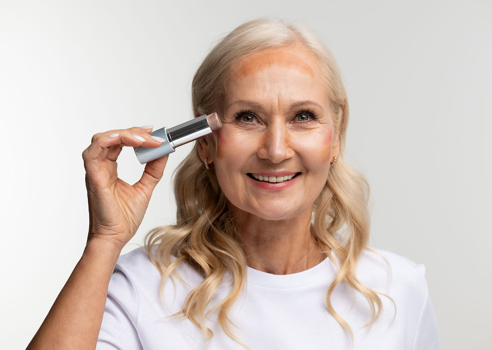 Smiling older woman with long blonde hair applies foundation or concealer stick to her cheek while wearing a white shirt, standing against a plain light background.