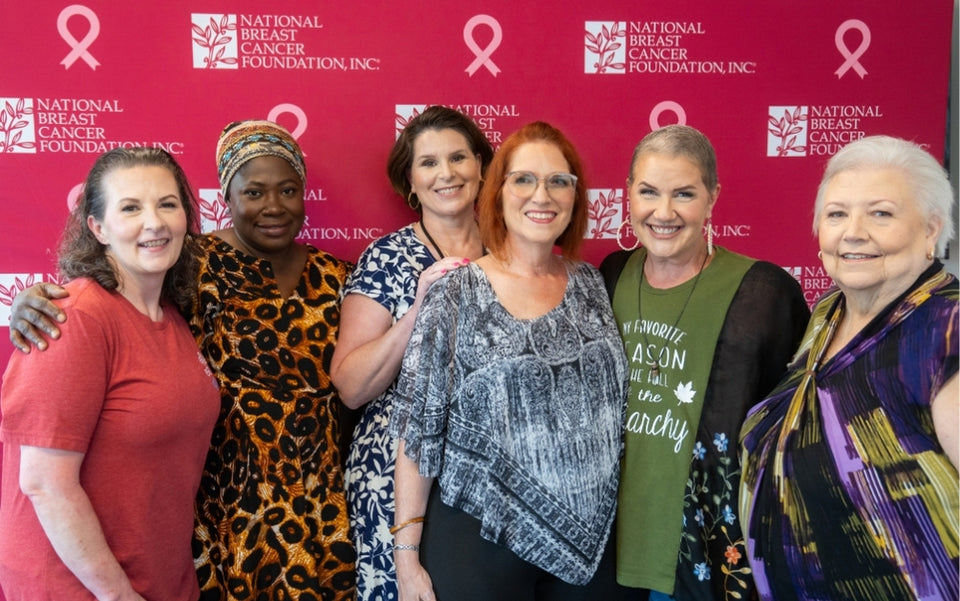 Six women stand together smiling in front of a National Breast Cancer Foundation, Inc. banner with pink ribbons, showing support and solidarity for breast cancer awareness.