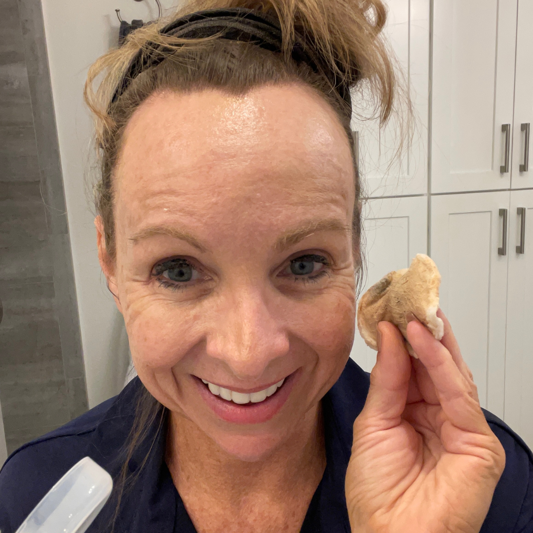 A smiling woman with a headband holds up a used cotton pad with visible makeup residue in one hand and a spray bottle in the other, standing in a bathroom with white cabinets in the background.