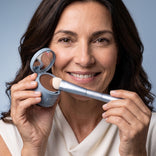 Eine lächelnde Frau hält das „Hairline Perfection Kit“ von PrimePrometics™ in der Hand, das eine Puderdose mit Spiegel und einen Applikatorpinsel enthält, und trägt damit gekonnt Haaransatzpuder auf, um graue Haare nahtlos abzudecken. Sie trägt ein weißes Oberteil und hat braunes Haar; im Hintergrund ist ein hellblauer Hintergrund zu sehen.
