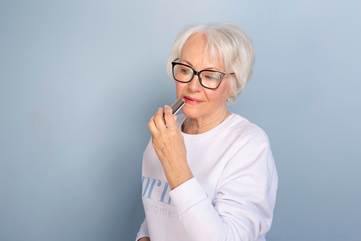 An older woman with short white hair and glasses applies lipstick while wearing a white sweatshirt, standing against a light blue background.