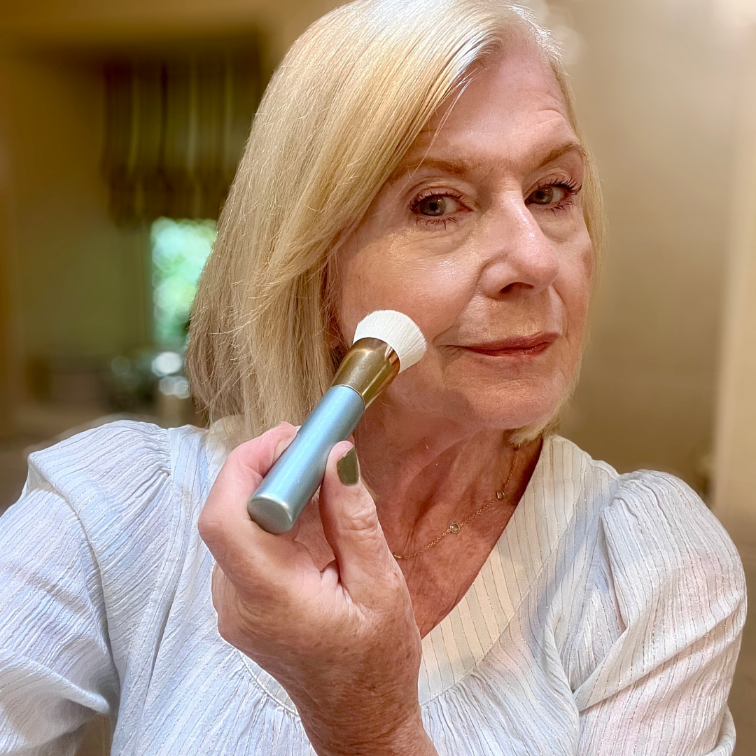An older woman with blonde hair applies makeup to her cheek using a brush. She is wearing a light-colored top and is standing indoors, possibly in a bathroom, with a slight smile on her face.