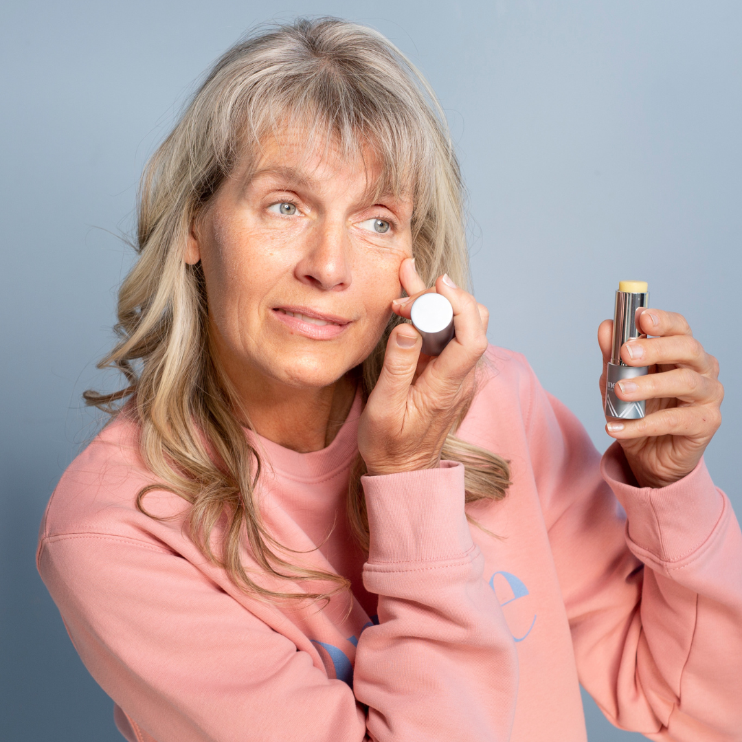 A woman with long blonde hair in a pink sweatshirt applies concealer under her right eye, holding a concealer stick in her left hand against a plain light blue background.