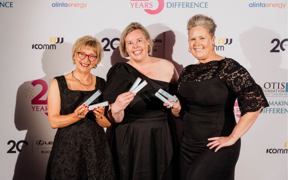 Three women dressed in black evening wear, smiling and holding awards, pose together in front of a step-and-repeat banner at an event.