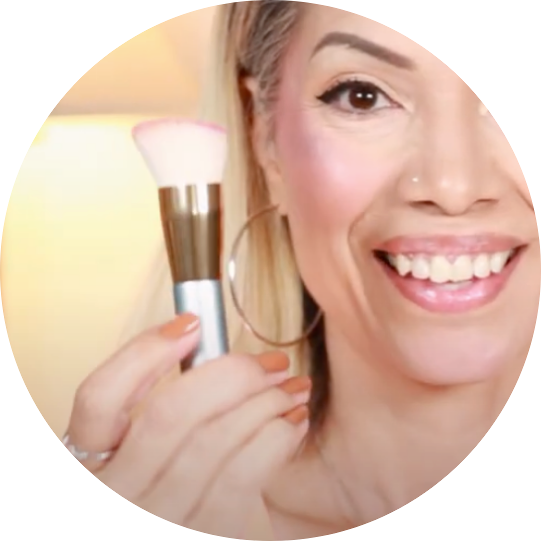 A smiling woman with light skin holds a makeup brush near her cheek, showing pink blush on her face. She has hoop earrings, a nose stud, and light-colored hair. The image is cropped in a circular frame.