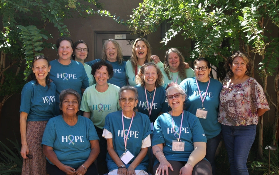 A group of thirteen women, some wearing teal HOPE t-shirts, smile and pose together outdoors in front of green foliage and a building. Most are standing; three are seated in front.