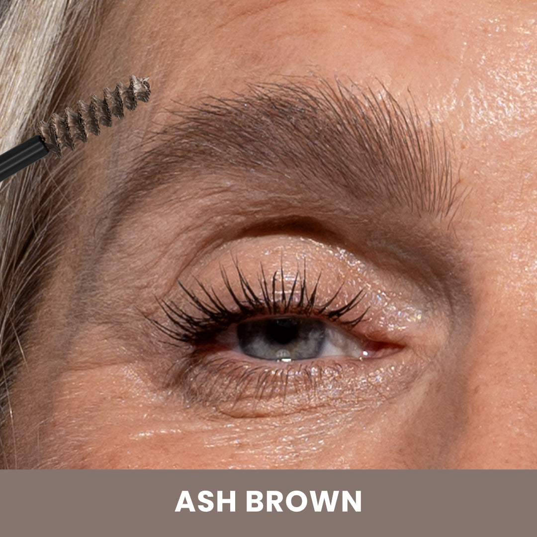 Close-up of an eye with grey lashes and a eyebrow brush touching a grey-brown eyebrow. The skin has natural texture. Text at the bottom reads ASH BROWN on a brown background.