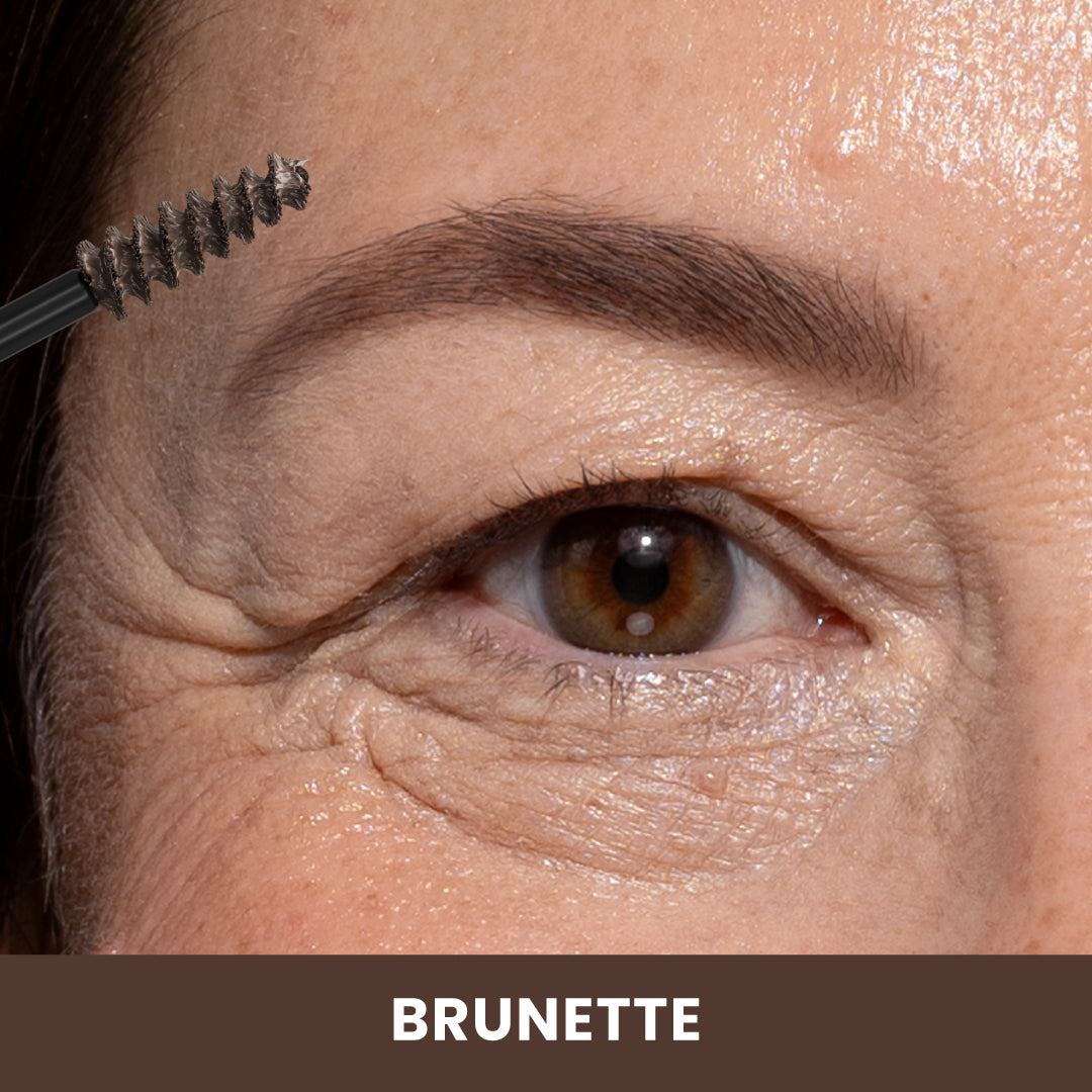 A close-up of a womans eye and eyebrow as a spoolie brush applies brow product. The image highlights a natural brunette brow color, with the word BRUNETTE at the bottom.