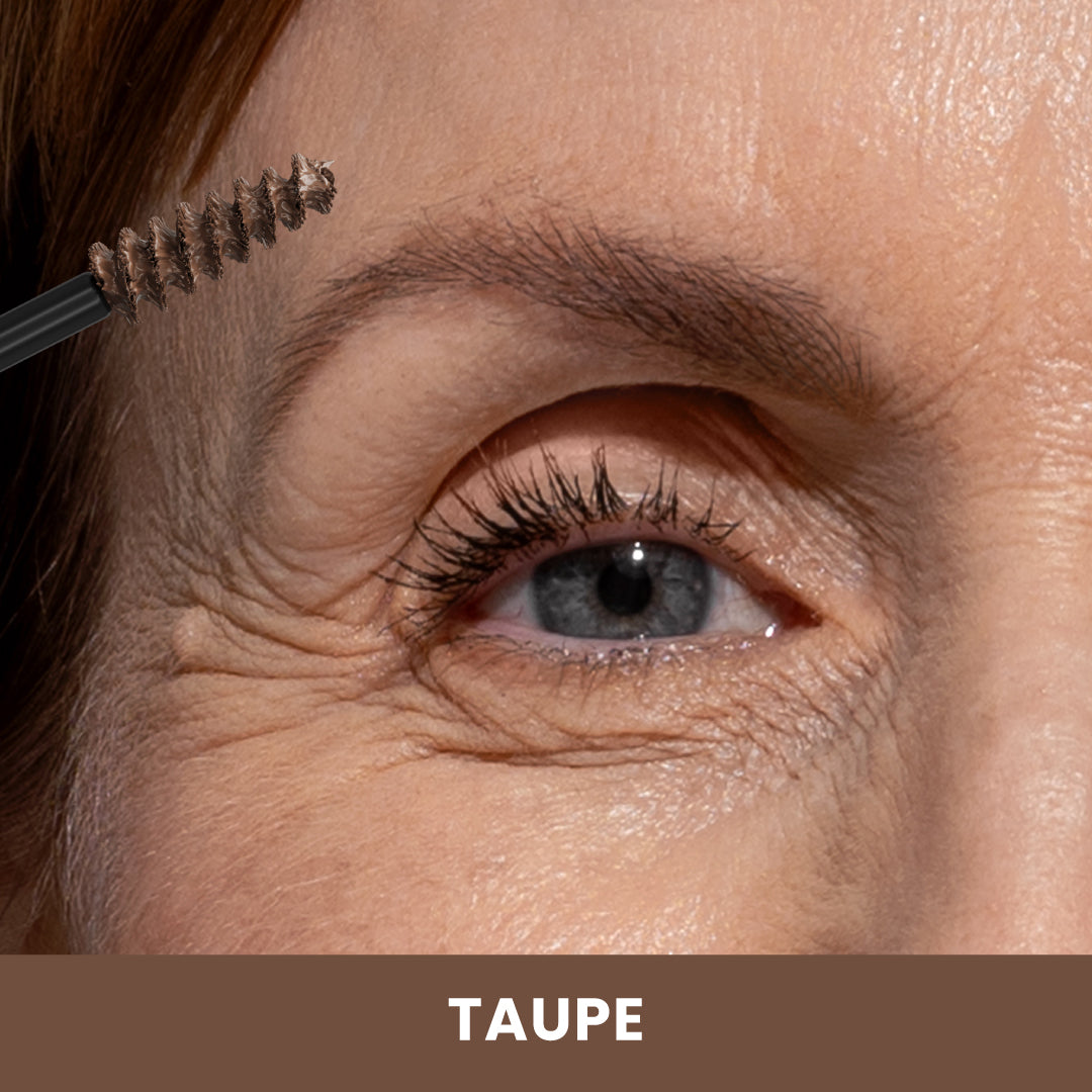 A close-up of a persons eye and eyebrow as they apply taupe-colored eyebrow gel with a spoolie brush. The skin shows natural wrinkles. The word TAUPE appears at the bottom of the image.