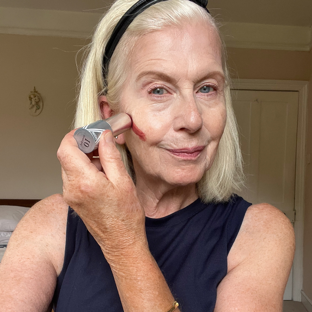 An older woman with blonde hair applies blush or makeup stick to her cheek while looking into the camera. She is wearing a sleeveless black top and a black headband, standing in a softly lit room.