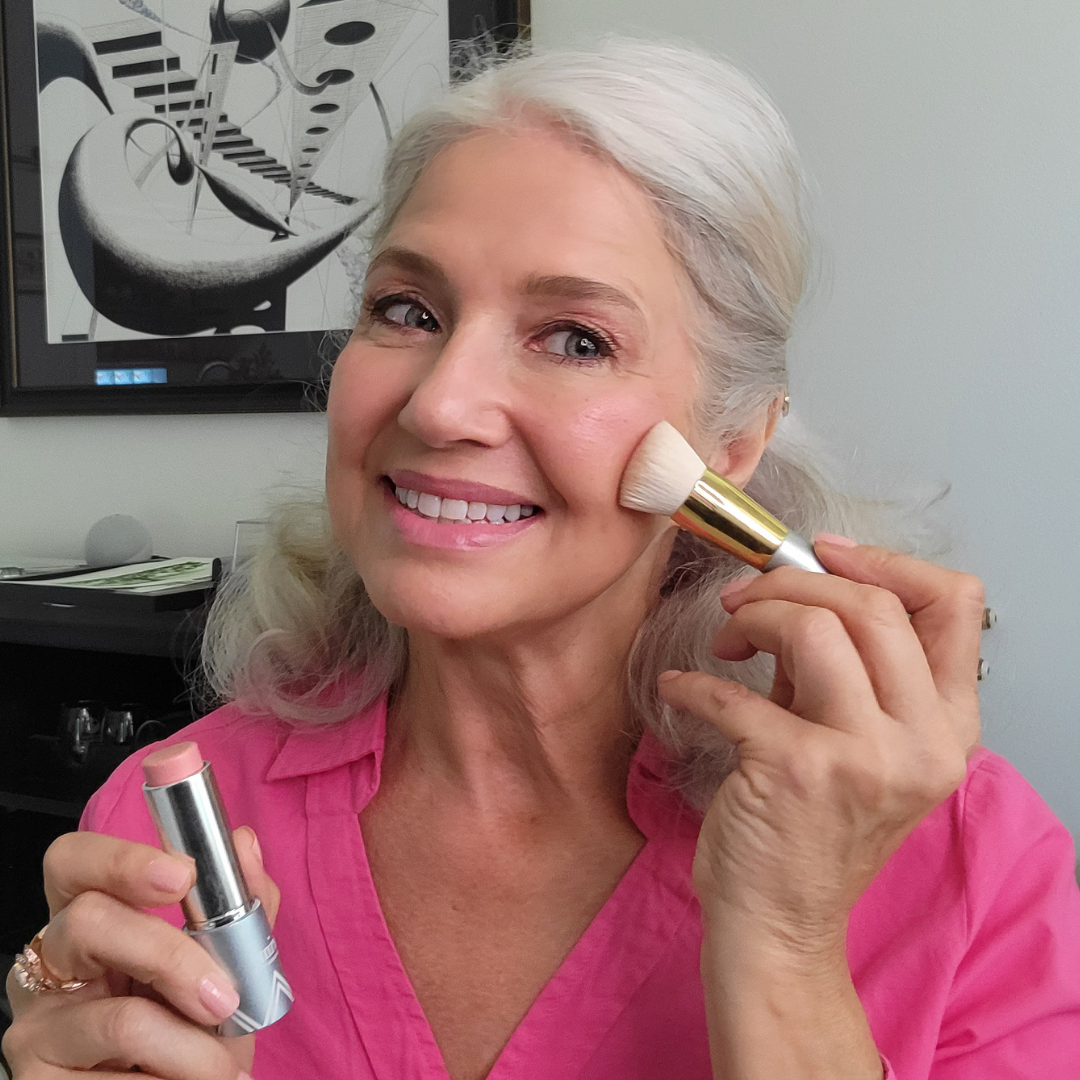 A smiling older woman with gray hair and a pink shirt applies blush to her cheek with a makeup brush, holding a stick of blush in her other hand. Abstract art hangs on the wall behind her.