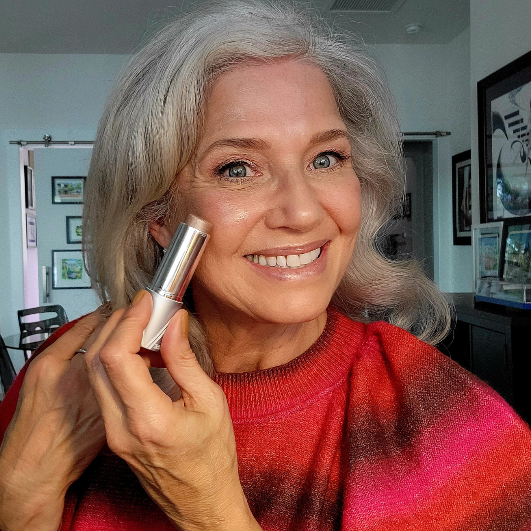 Smiling older woman with gray hair applies makeup to her cheek with a stick applicator. She wears a red sweater and is indoors, with framed pictures hanging on the walls behind her.