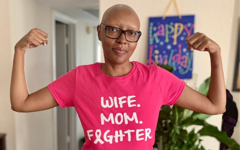 A woman with glasses and a shaved head flexes her arms, wearing a pink shirt that reads Wife. Mom. Fighter. Behind her is a colorful Happy Birthday sign and green indoor plants.