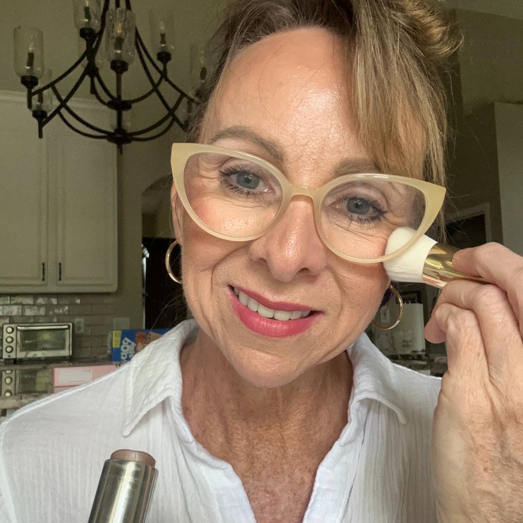 A woman with light brown hair and large glasses is smiling while applying makeup with a brush to her cheek. She is wearing a white top and is in a kitchen with light-colored cabinets and a black chandelier in the background.