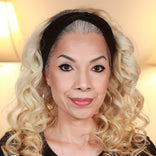A woman with long, curly blonde hair, wearing a black headband, hoop earrings, and a black top, smiles softly at the camera in a warmly lit indoor setting.