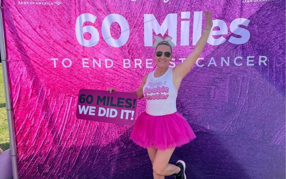 A smiling woman in a pink tutu and sunglasses poses in front of a purple banner that reads 60 Miles to End Breast Cancer. She holds a sign saying 60 MILES! WE DID IT! and has one arm raised in celebration.
