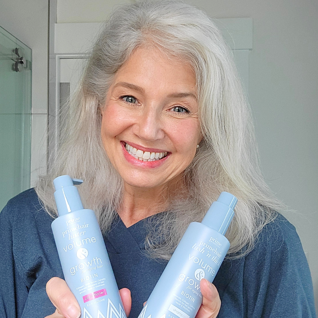 Smiling woman with long gray hair holds two blue bottles of hair care products labeled Volume and Growth in a bright bathroom setting.