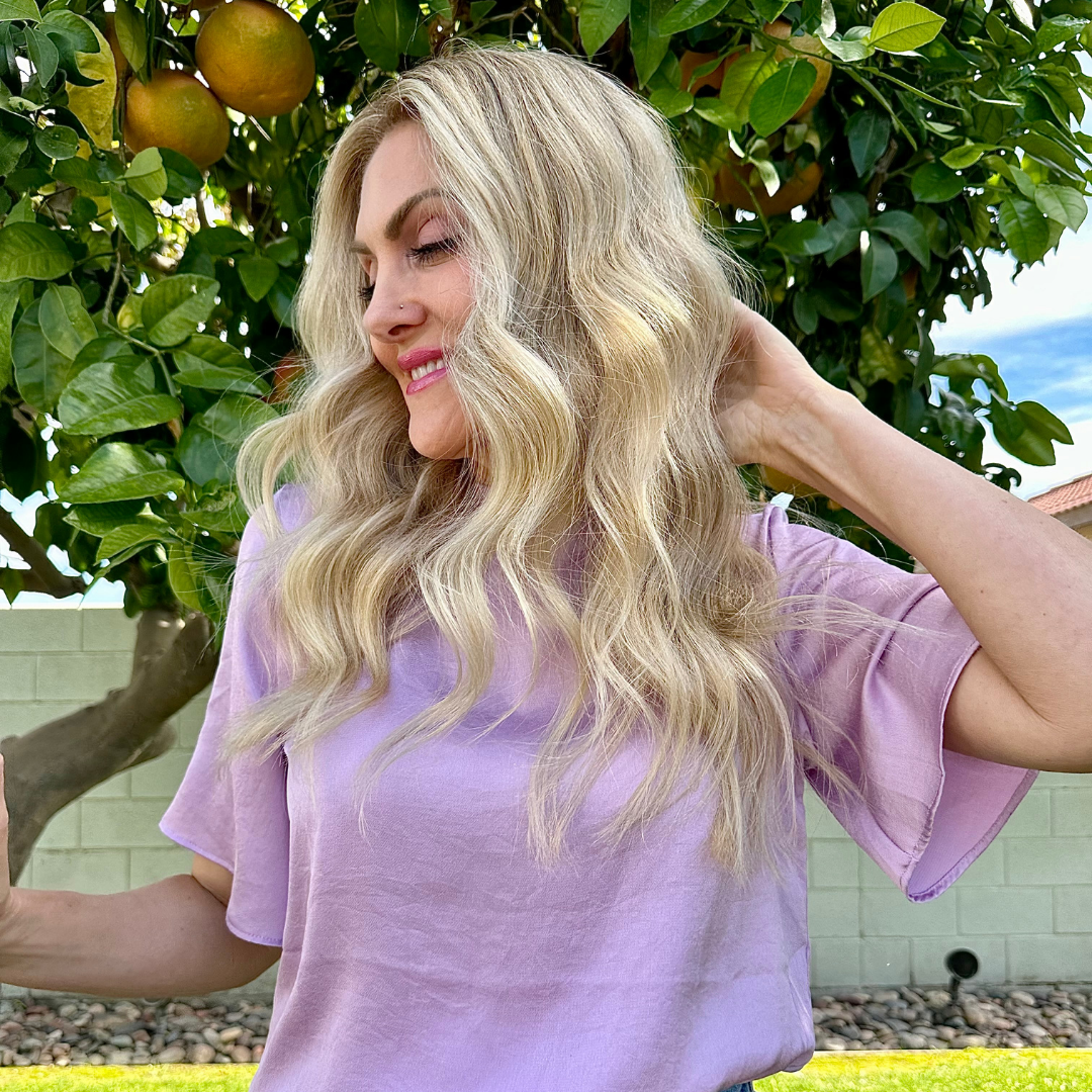 A woman with long, wavy blonde hair wearing a light purple shirt smiles while standing outdoors near a leafy tree with large orange fruits.
