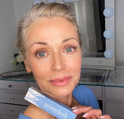 A woman with light hair and blue eyes holds a blue Primelash mascara box, smiling in front of a vanity mirror with round lights.