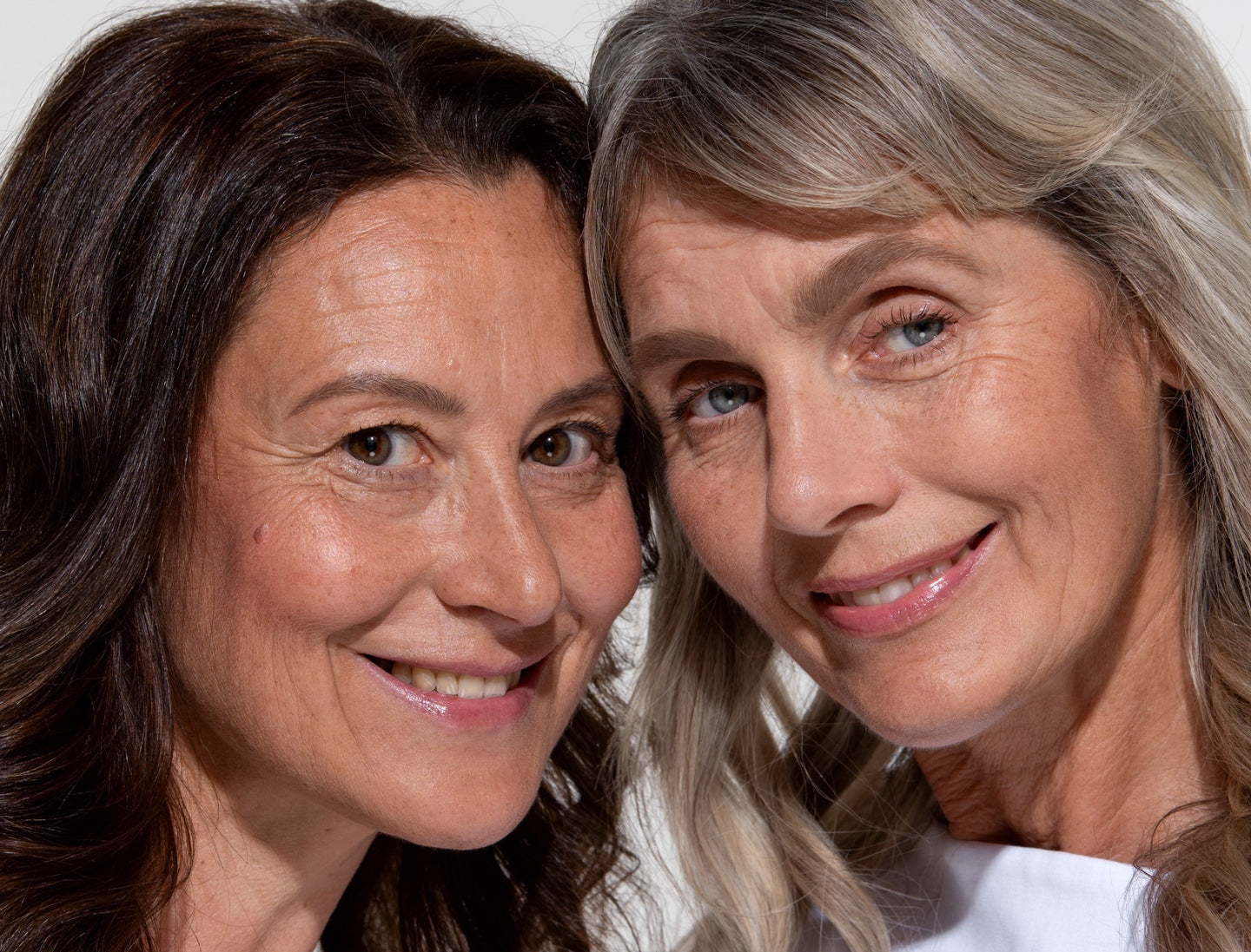 Two women with long hair, one brunette and one with gray-blonde hair, stand close together and smile warmly at the camera against a neutral background.