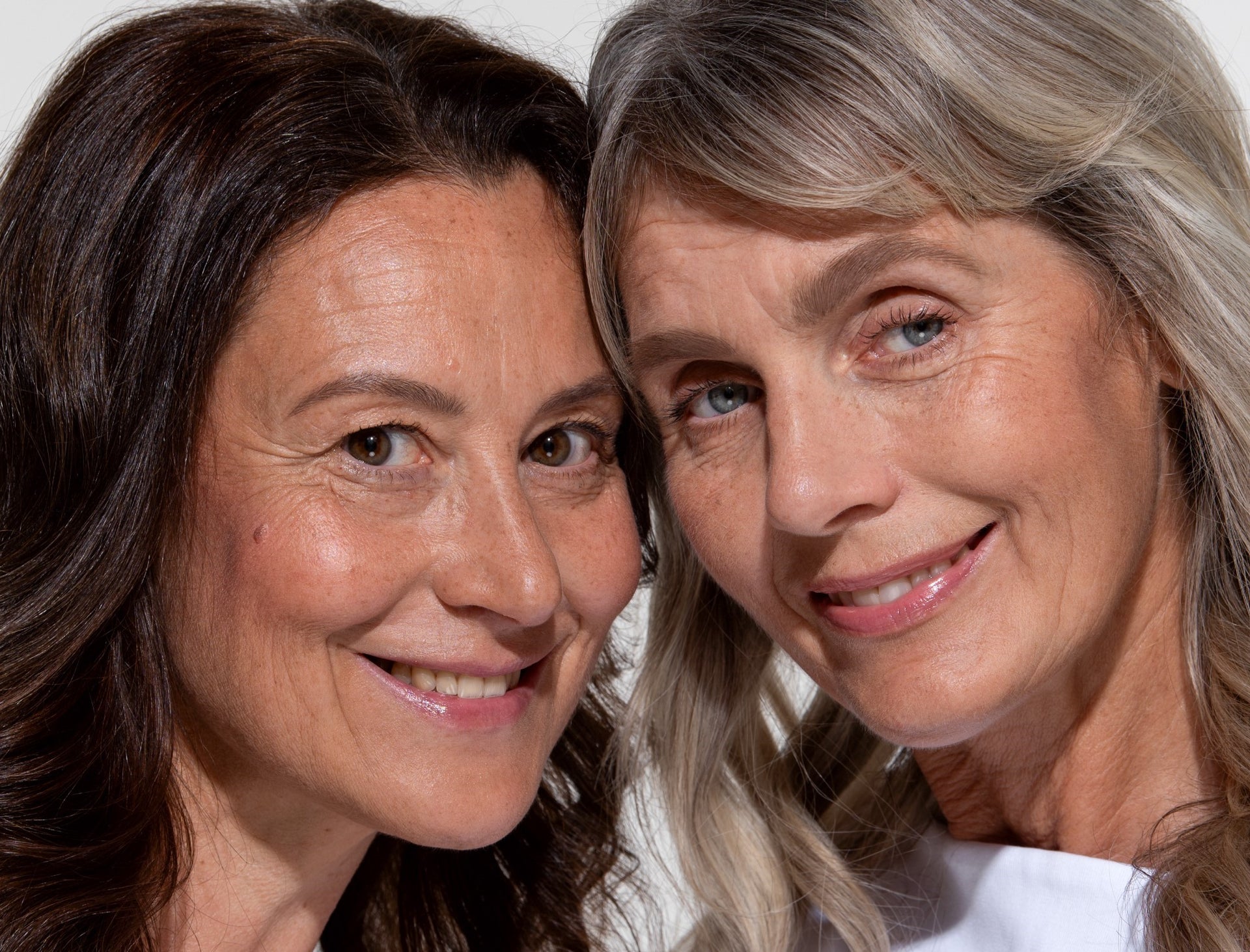 Two women with long hair, one brunette and one with gray-blonde hair, stand close together and smile warmly at the camera against a neutral background.