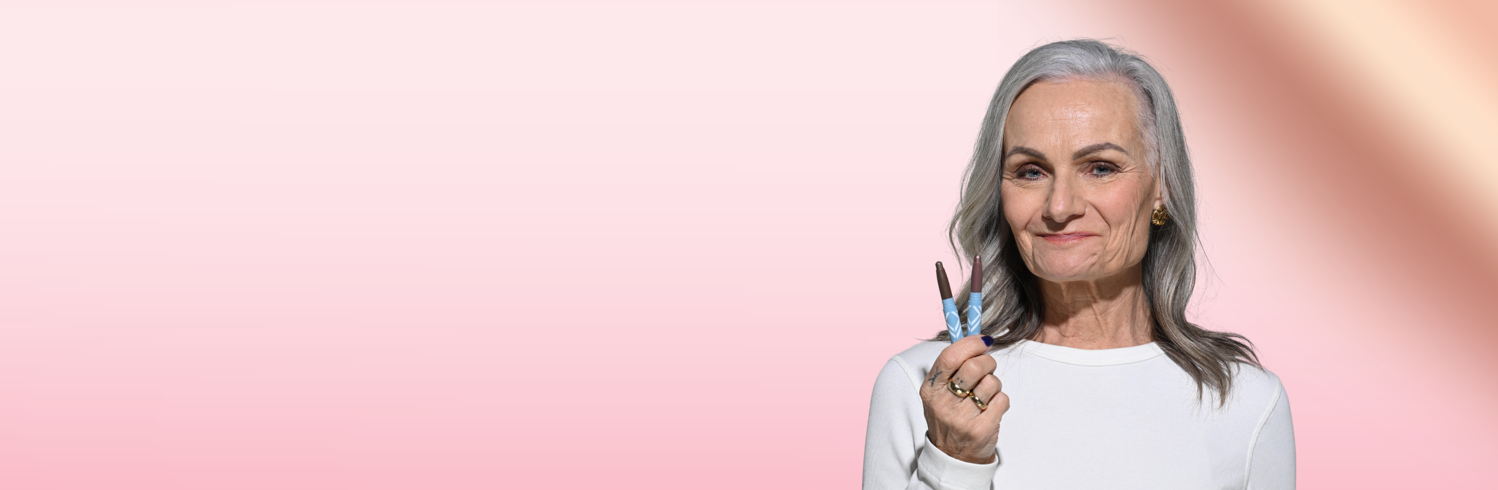 An older woman with long gray hair and a white top holds up a lipstick and smiles confidently against a pink gradient background.