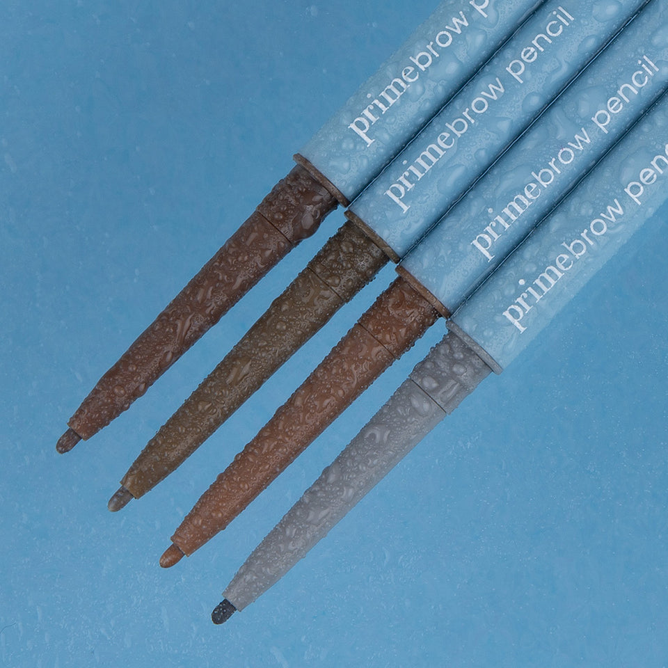 Four eyebrow pencils labeled primebrow pencil are arranged diagonally on a textured blue surface. Each pencil has a different shade, ranging from dark brown to light grey.