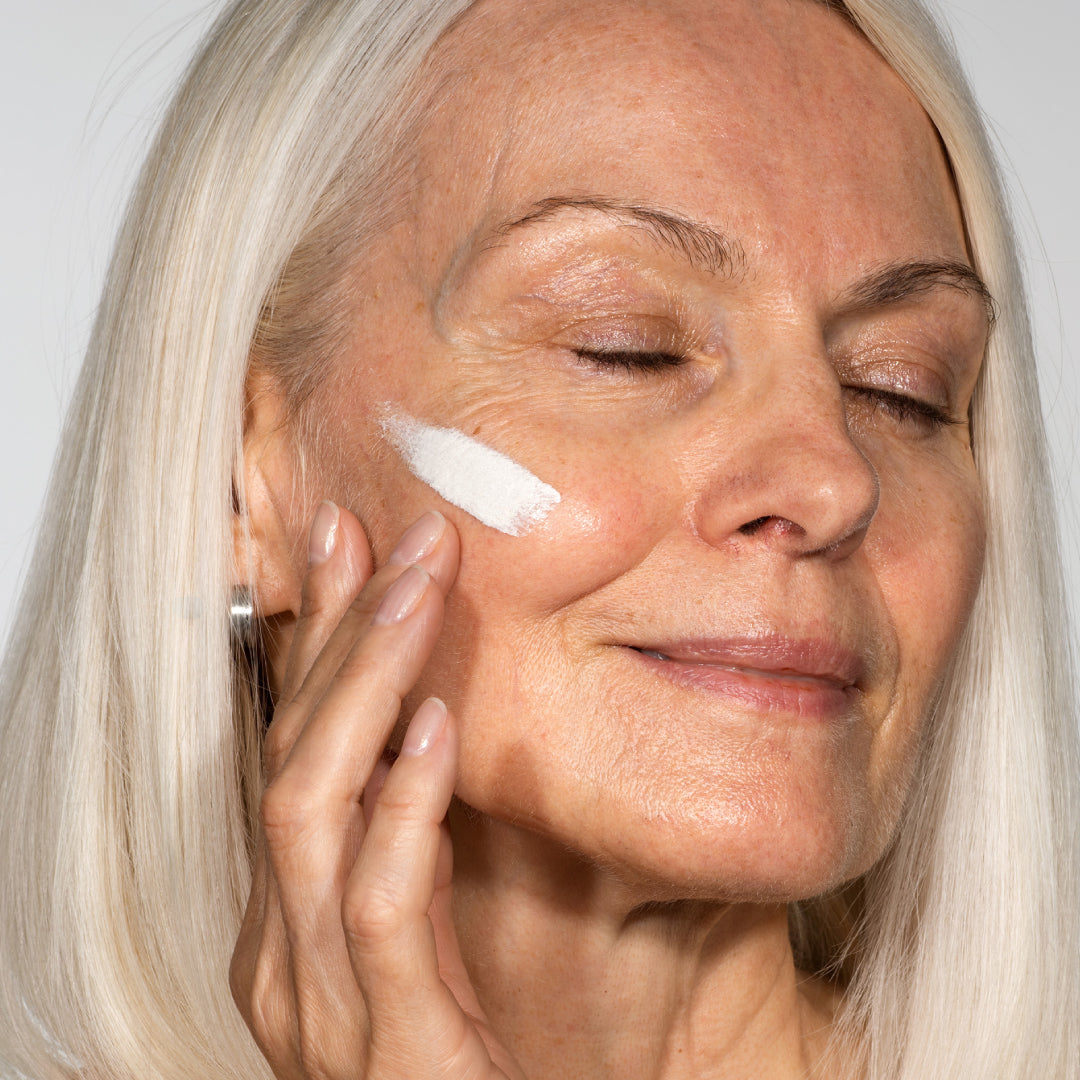An older woman with long white hair and closed eyes applies a streak of white cream to her cheek, gently massaging it in with her fingers. She appears relaxed and content.