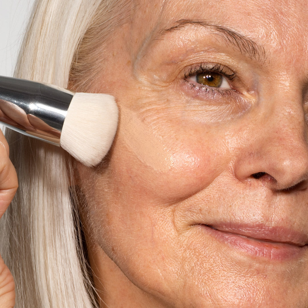 A close-up of an older woman with light skin and gray hair applying foundation to her cheek with a makeup brush, highlighting smooth and healthy skin.