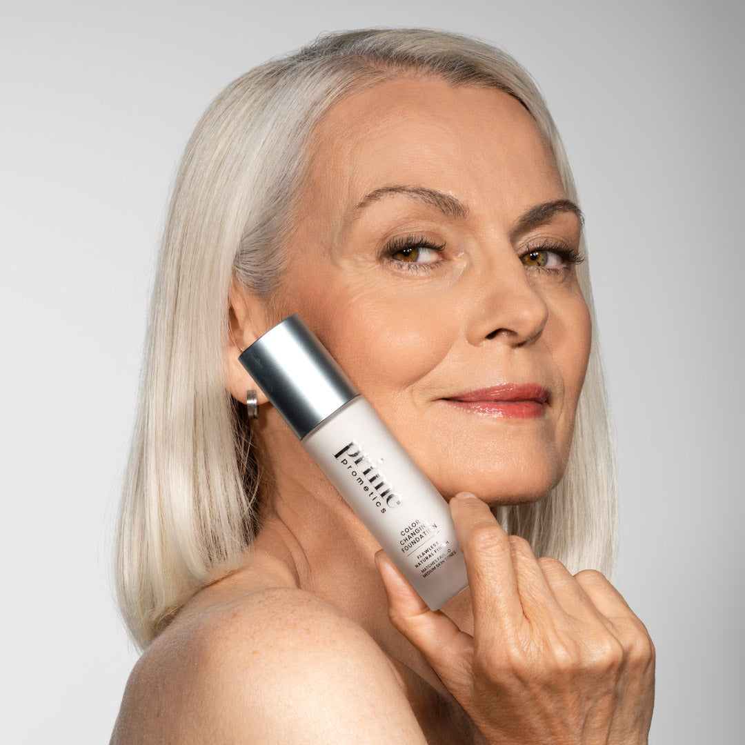 A smiling older woman with long, straight gray hair holds a silver-capped bottle of skin care product near her cheek against a plain light background.