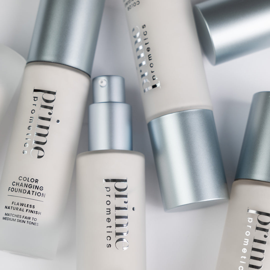 A close-up of several silver-capped bottles labeled prime prometics color changing foundation, arranged on a white surface. Some bottles are lying down, while one is upright with the pump exposed.